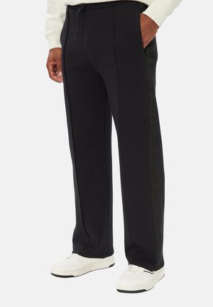 Persona che indossa pantaloni neri a gamba dritta con strisce laterali e sneakers bianche, in piedi con una mano in tasca.
