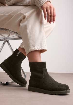 Suede dark green ankle boots with a round toe and side zipper, paired with light beige, cuffed trousers, on a smooth surface.