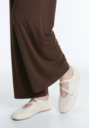 Personne portant un pantalon marron à jambes larges et des chaussures style Mary Jane crème avec double brides, debout sur une surface blanche.