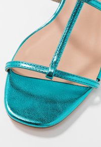 Turkoosi metallinhohtoinen sandaali litteällä T-nauha -designilla. Sisältää sileän, kiiltävän pinnan ja minimalistiset nauhat. Suljettu varvastyylinen.