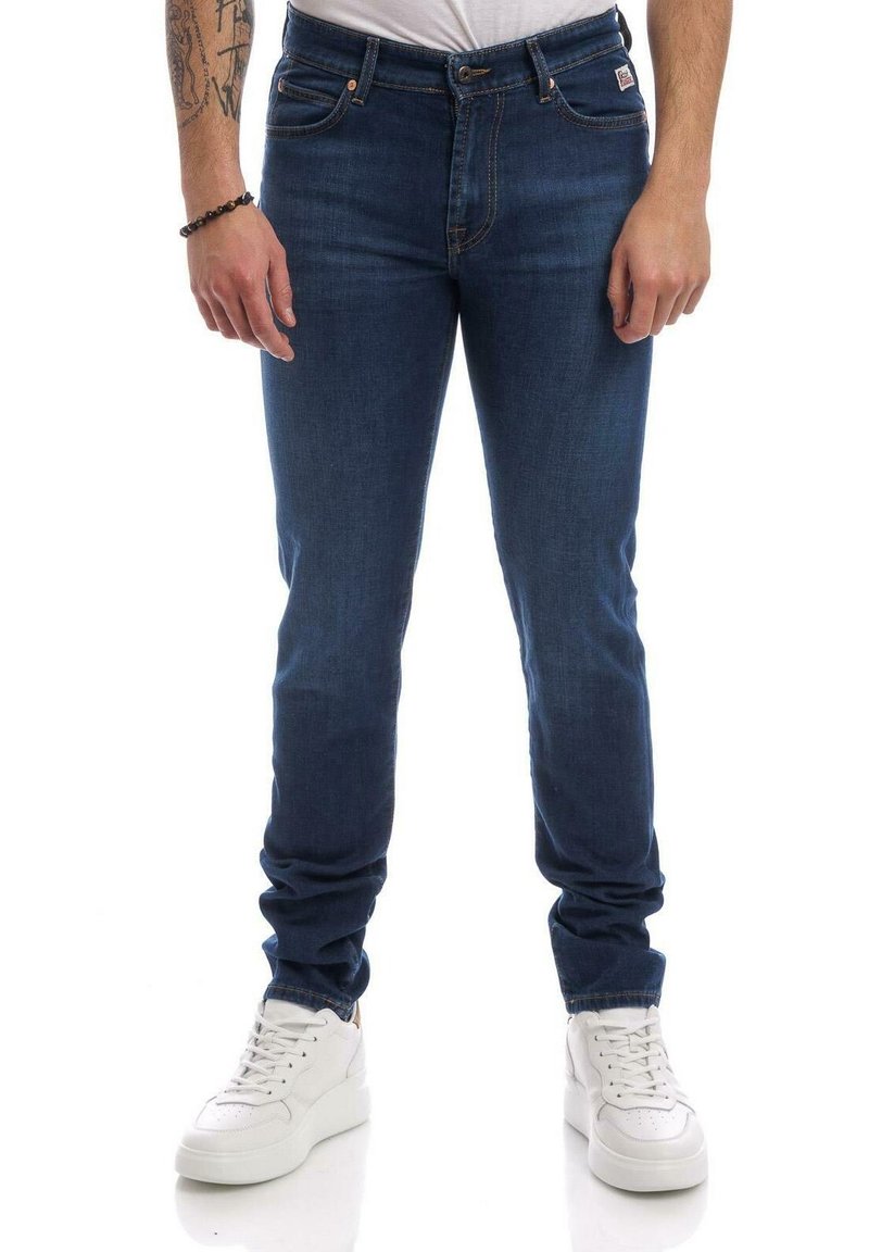 Tmavě modré džíny se slim střihem, světlým prošíváním, pěti kapsami a zapínáním na knoflíky, doplněné bílými teniskami.