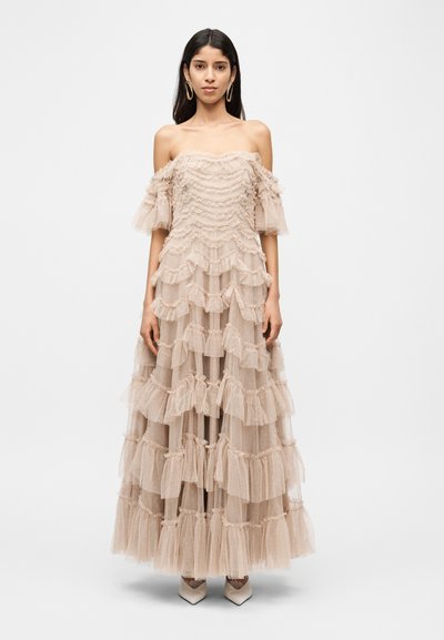 Robe en tulle beige à épaules dénudées avec des volants superposés, un corsage texturé et des manches évasées. Portée avec des talons à bouts pointus.