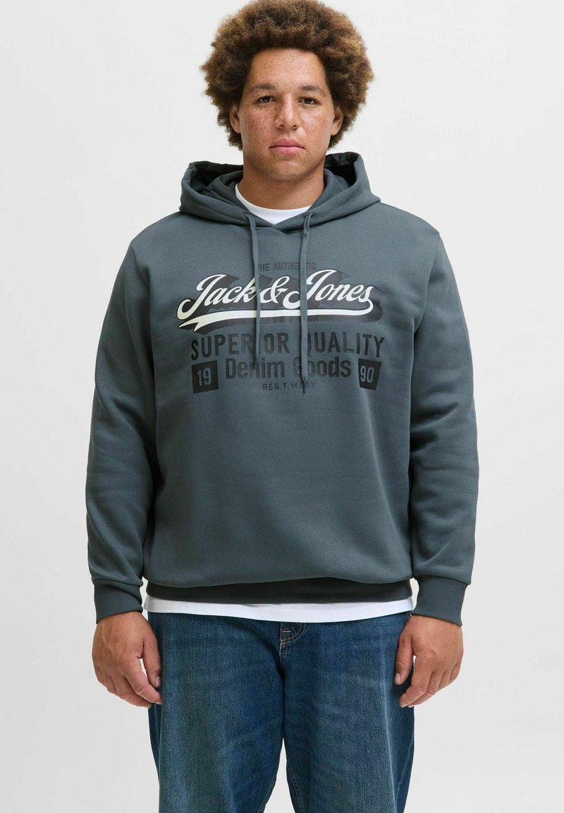 Grauer Kapuzenpullover mit Kordelzug, der ein großes Logo und Text in Weiß und Schwarz zeigt. Über einem weißen Shirt getragen und mit blauen Jeans kombiniert.