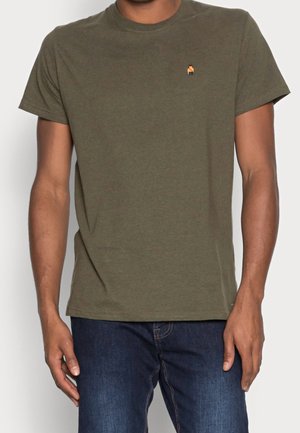 Mann trägt ein olivgrünes Kurzarm-T-Shirt mit einem kleinen orange-schwarzen Logo auf der linken Brust, kombiniert mit dunkelblauen Jeans.