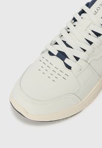 Baskets gris clair en cuir lisse avec des lacets blancs, des accents bleu marine, un orteil perforé et une semelle en caoutchouc avec un grip texturé.
