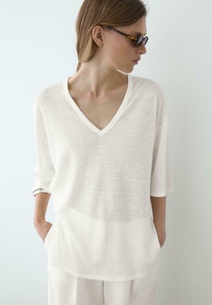 Frau trägt übergroßes weißes V-Ausschnitt-Top und helle Hose, mit Händen in den Taschen und Schildpatt-Sonnenbrille, schaut zur Seite.