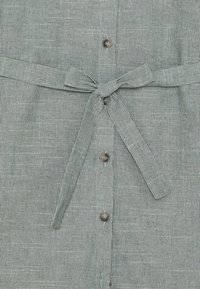 Pionowa, szaro-prążkowana tkanina z paskiem w talii związanym w kokardę, umieszczonym nad czterema brązowymi guzikami na środku.