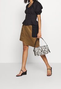 Schwarzes Oberteil mit Puffärmeln aus strukturiertem Stoff, kombiniert mit einem braunen Rock mit einer schwarzen aufgesetzten Tasche. Hält eine Handtasche mit Zebra-Print.