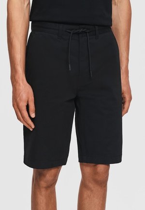 Mężczyzna w czarnych, sięgających do kolan szortach ze sznurkiem w talii i bocznymi kieszeniami, stojący na tle jednolitego jasnego tła.