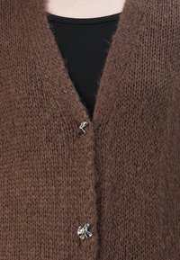 Brun strikket cardigan med to sølv abstrakte knapper over en svart skjorte, nærbilde av brystområdet.