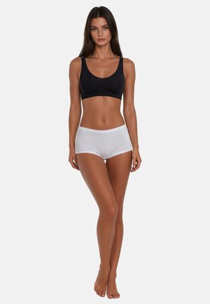 Woman standing barefoot wearing a black sports bra and white boyshort underwear against a plain light background.