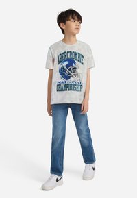 Grafiska T-krekls ar zilu futbola aizsardzību un tekstu "Abercrombie Nacionālā čempionāta", apvienots ar zilām džinsām un baltām sporta kedām.