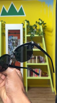 Juodi akiniai nuo saulės su tamsiais lęšiais, kampuotomis kojelėmis ir blizgiu paviršiumi. Ant šventyklos yra subtilus logotipas. Laikomi rankoje priešais spalvingą foną.