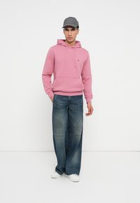 Różowa bluza z kapturem, z kieszenią z przodu i sznurkiem do regulacji. Jasnoniebieskie, szerokie dżinsy o wyblakłym odcieniu i detalach w szyciu. Szara czapka i białe trampki.