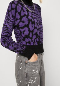 Pull imprimé léopard violet et noir avec un ourlet côtelé noir et un col roulé, porté avec un jean gris éclaboussé de peinture blanche.