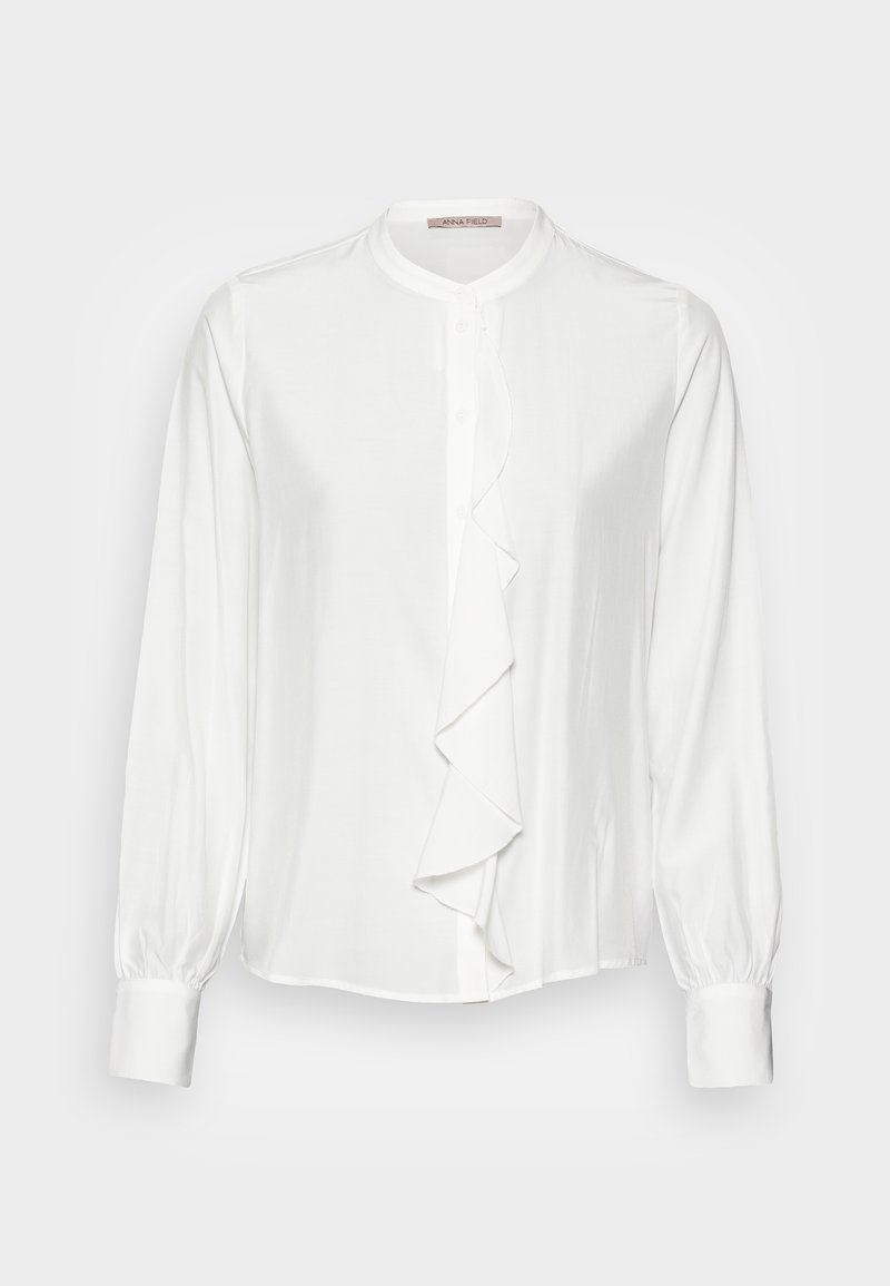 Blusa bianca con colletto alto, chiusura frontale con bottoni e dettaglio di rouches a cascata lungo la patta. Maniche lunghe e arricciate con polsini.