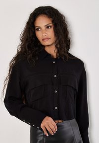 Czarna krótka koszula z guzikami z dwoma przednimi kieszeniami, srebrnymi guzikami i kołnierzykiem. Długie rękawy z guzikami na mankietach.
