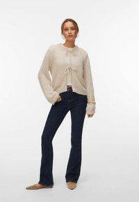 Femme portant un cardigan beige en tissu duveteux avec des nœuds attachés, un jean évasé bleu foncé, des ballerines pointues marron clair et des boucles d'oreilles créoles, debout devant un fond blanc.