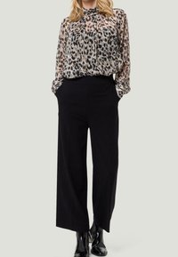 Blusa in tessuto trasparente con stampa leopardata, abbinata a pantaloni neri a gamba larga. La blusa presenta un colletto arricciato e maniche lunghe. Stivaletti neri alla caviglia.