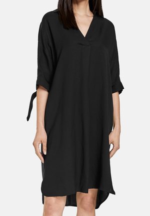 Femme portant une robe noire ample jusqu'aux genoux avec un col en V et des manches 3/4 nouées aux coudes, posant devant un fond uni.