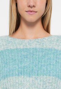 Mladá žena s rovnými blond vlasy, oblečená v modro-bílé pletené svetr, ukazuje rty a oblast krku.