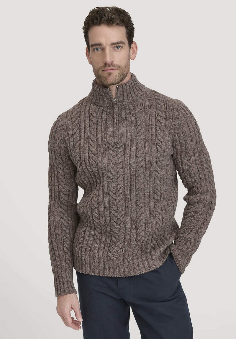 Brauner Strickpullover mit hohem Kragen und halbem Reißverschluss, der mit strukturierten Zopfmustern versehen ist. Über einer marineblauen Hose getragen.