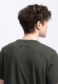 Maglietta in cotone verde scuro con collo a giro, caratterizzata da un sottile logo a forbice grigio sul retro, vicino al collo. Texture liscia.