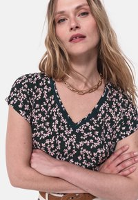 Top a stampa floreale di colore blu scuro con fiori rosa. Presenta maniche corte, scollo a V e un bordo in pizzo decorativo lungo lo scollo.