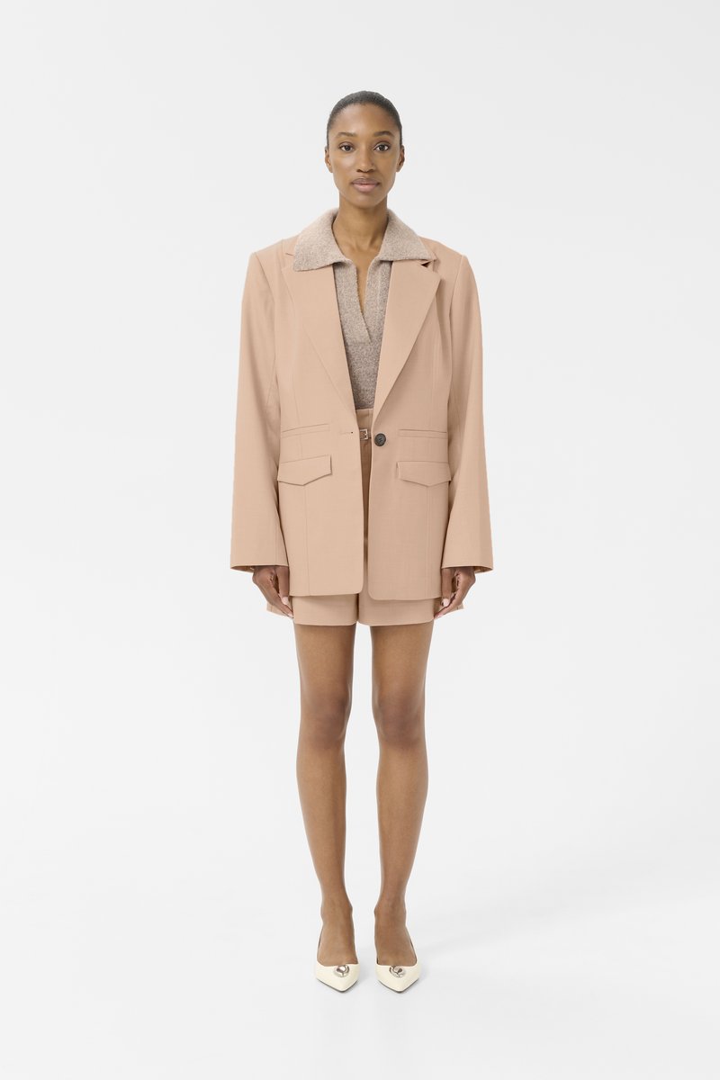 Světlý béžový blazer s měkkým límcem, doplněný pleteným topem a odpovídajícími šortkami. Modelka má na sobě krémové lodičky. Jednoduchý, dobře střižený design.