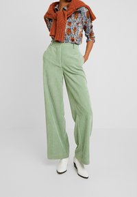 Pantalones de pana verde de pierna ancha con una superficie texturizada, combinados con una blusa estampada y un suéter de punto naranja atado alrededor de los hombros.
