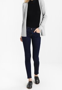 Šedý cardigan přes černý žebrovaný top, v kombinaci s tmavě modrými skinny džíny a černými šněrovacími botami. Hladké textury, štíhlá silueta.