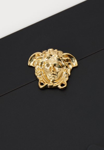 Czarna koperta z emblemą złotej głowy Meduzy jako zamknięciem, z wyraźnie zaznaczonymi rysami twarzy i falującymi akcentami włosów.