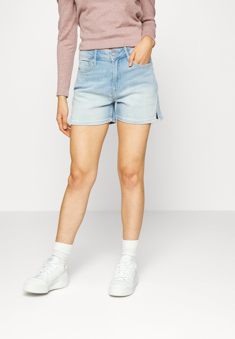 Ljust blå denimshorts med avslappnad passform, fållade kanter och sidosprund. Kombinerat med vita sneakers och långärmad tröja med ribbat mönster.