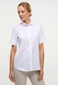 Camicia bianca a maniche corte con bottoni, realizzata in tessuto liscio, caratterizzata da un colletto classico e una vestibilità sagomata con un leggero lucido.