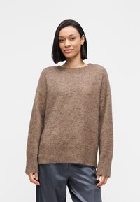 Καφέ πλεκτό πουλόβερ από μαλακό ύφασμα με άνετη εφαρμογή, στρογγυλή λαιμόκοψη και ριμπ μανσέτες, συνδυασμένο με σκουρόχρωμο γκρι παντελόνι.