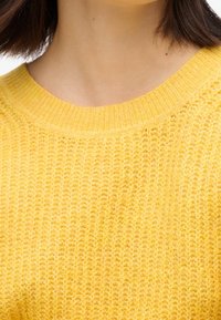 Maglione giallo a maglia con scollatura a coste, caratterizzato da un motivo testurizzato e un tessuto morbido. La vista ravvicinata mette in evidenza i dettagli della trama.