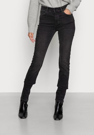 Person, die hoch taillierte schwarze Skinny-Jeans, einen grauen Langarmpullover und schwarze Stiefeletten trägt, steht mit einer Hand in der Tasche.