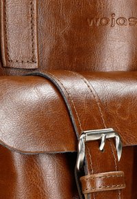 Bruine leren tas met een gestructureerde afwerking, voorzien van een zilveren metalen gesp en stiksels. Het ontwerp omvat een flap en een verstelbare schouderband.