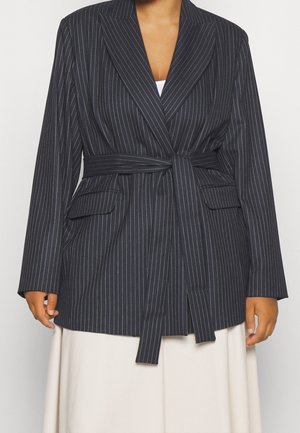 Vrouw draagt een donker colbert met fijne strepen en een tailleband over een witte top en een crème rok, getoond van schouders tot halverwege de dij.
