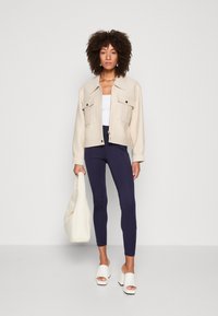Beige korte jas met twee voorzakken, witte tanktop, donkerblauwe legging en witte blokhak sandalen. Houdt een crèmekleurige tote bag vast.