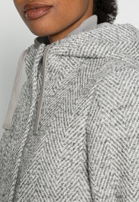 Szara bluza z kapturem w jodełkę wykonana z miękkiego, teksturowanego materiału; posiada sznurek ściągający oraz prążkowany dekolt.