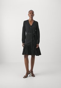 Black button-up dress with long sleeves, tie waist, and a flared skirt featuring ruffles. Smooth fabric, minimalistic design, knee-length.