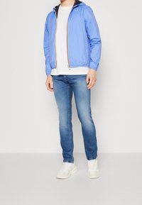 Ljusblå zip-up-jacka med luva, med svart innerfoder som kontrast, kombinerad med vit skjorta och blå jeans. Sneakers fullbordar looken.