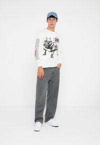 Wit T-shirt met lange mouwen, voorzien van een buldogafbeelding en een zwart netpatroon op de mouw, gecombineerd met grijze jeans en witte sneakers.