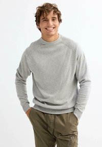 Jersey de punto gris con cuello y puños acanalados, llevado sobre pantalones verde oliva. El modelo se encuentra de pie con las manos en los bolsillos, sonriendo.