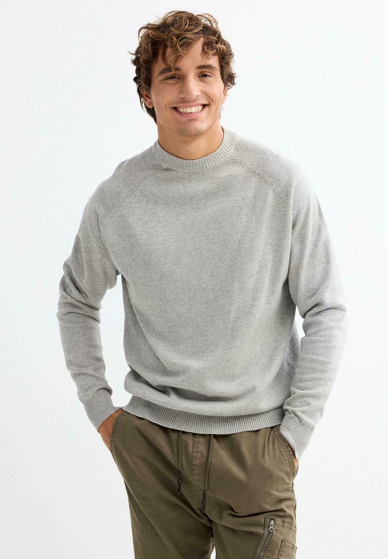 Jersey de punto gris con cuello y puños acanalados, llevado sobre pantalones verde oliva. El modelo se encuentra de pie con las manos en los bolsillos, sonriendo.