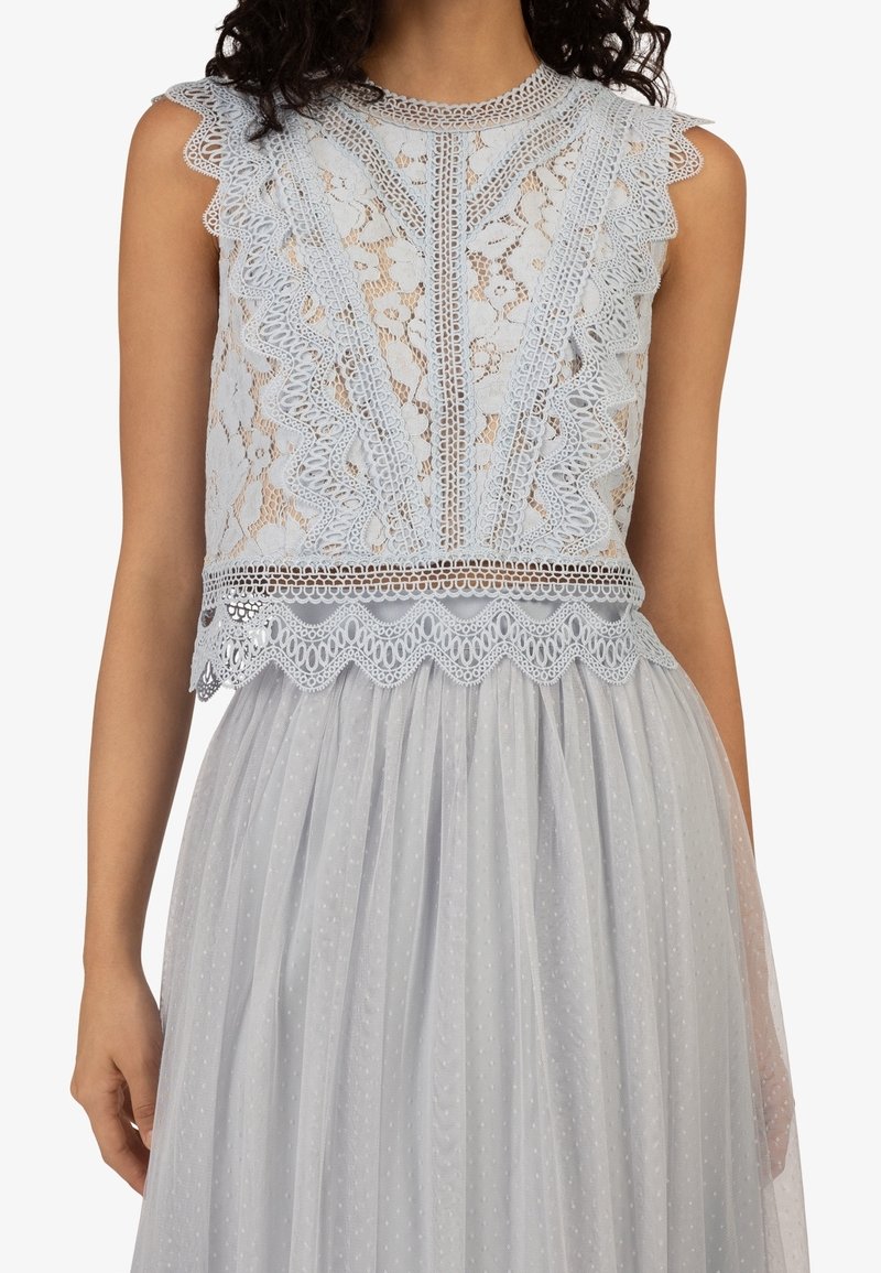 Femme portant un haut en dentelle gris clair sans manches avec des bords festonnés et une jupe plissée transparente avec un motif subtil à pois.