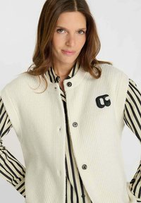 Canottiera bianca a costine con colletto arrotondato, bottoni neri e un logo nero; abbinata a una camicia a righe con righe nere e bianche.