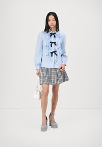 Light blue long-sleeve blouse with ruffle accents and black bows. Paired with a plaid skirt and grey heeled shoes, holding a white handbag.