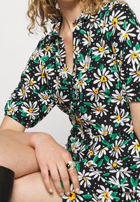 Blumenkleid mit Margeriten in Weiß, Gelb und Grün auf schwarzem Hintergrund; kurze Ärmel und eine Knopfleiste.